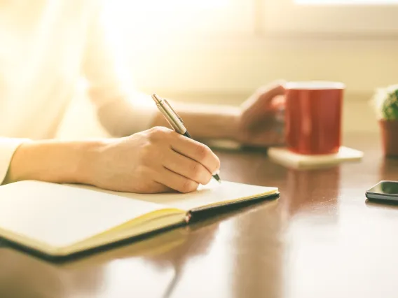 Person writing with a pencil in a notebook with a red cup next to them