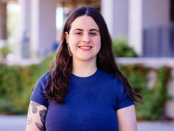  A smiling woman with long, dark wavy hair stands outdoors in front of a softly blurred backdrop. She wears a fitted short-sleeve blue shirt, dangling earrings, and has a floral tattoo on their left upper arm. 