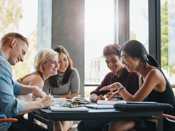 Five college aged people sit around a table laughing with notebooks, computers, and other office supplies.
