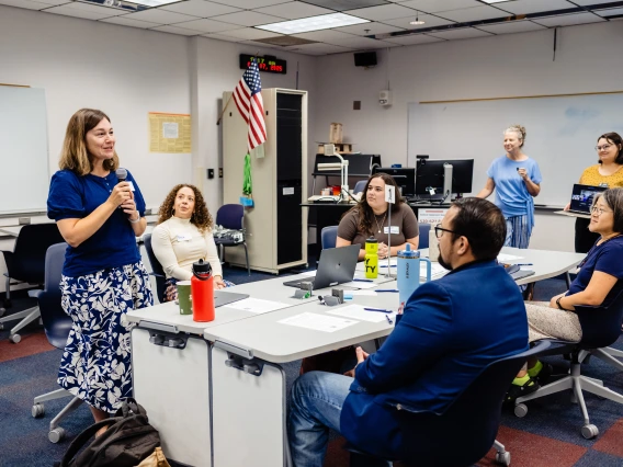 A group of adults in a classroom meeting. A woman stands speaking with a microphone, while others are seated at a table with laptops, one displaying remote attendees.