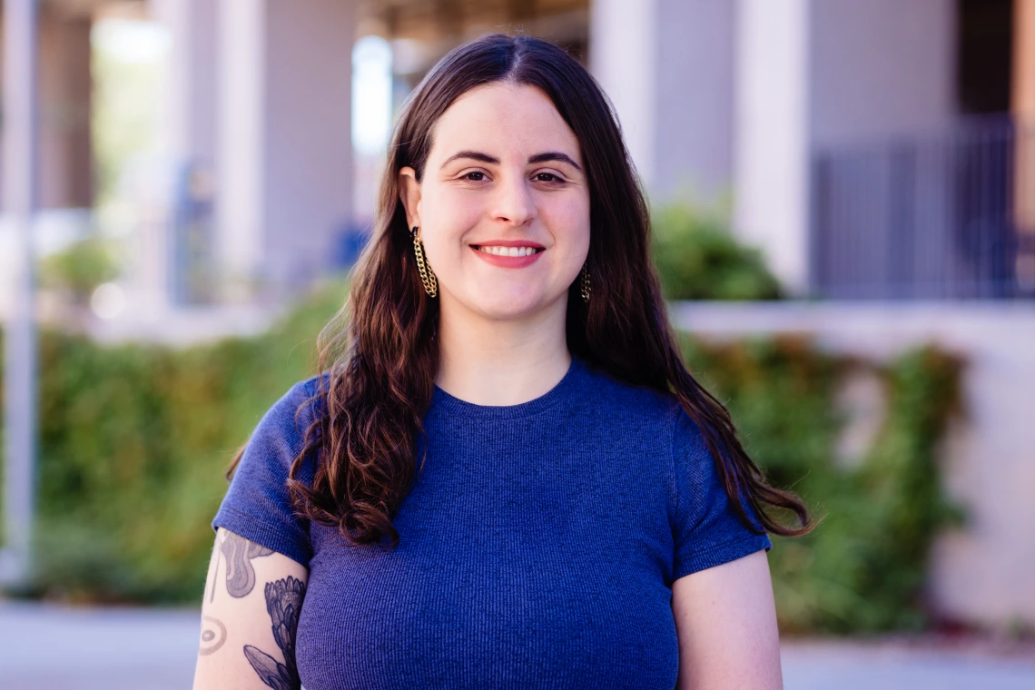  A smiling woman with long, dark wavy hair stands outdoors in front of a softly blurred backdrop. She wears a fitted short-sleeve blue shirt, dangling earrings, and has a floral tattoo on their left upper arm. 