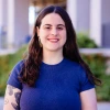  A smiling woman with long, dark wavy hair stands outdoors in front of a softly blurred backdrop. She wears a fitted short-sleeve blue shirt, dangling earrings, and has a floral tattoo on their left upper arm. 